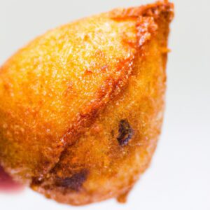 Fotografia de uma coxinha da asa assada em uma fritadeira elétrica. A coxinha é dourada e crocante, com a pele torrada e suculenta. O fundo é branco, com a fritadeira elétrica visível no canto inferior direito.