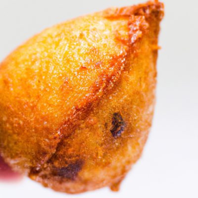 Fotografia de uma coxinha da asa assada em uma fritadeira elétrica. A coxinha é dourada e crocante, com a pele torrada e suculenta. O fundo é branco, com a fritadeira elétrica visível no canto inferior direito.