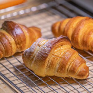 Imagem de croissants folheados dourados e crocantes cozidos na Airfryer Lidl. Os croissants têm uma textura escamosa e um interior macio e amanteigado. Eles são servidos em uma cesta de vime com uma xícara de café ao lado.