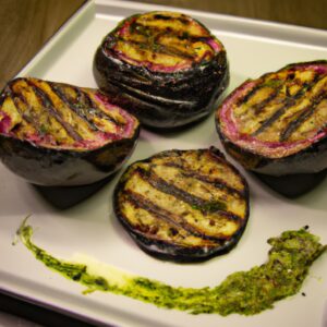 Uma vista superior de um medalhão de berinjela fatiado em rodelas grossas e empanado, coberto com pesto verde. O prato está disposto em um prato branco com uma guarnição de folhas de manjericão fresco.