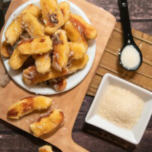 Imagem de palitinhos de banana crocantes e dourados em uma frigideira, polvilhados com queijo parmesão ralado. Os palitinhos são finos e cortados na diagonal, criando uma apresentação atraente.
