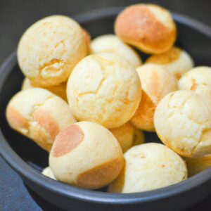 Foto de vários pães de queijo quentes e dourados em uma airfryer. Eles são recheados com catupiry derretido e polvilhados com orégano. A imagem também mostra os ingredientes usados para fazer o pão de queijo, como polvilho azedo, queijo parmesão e leite.