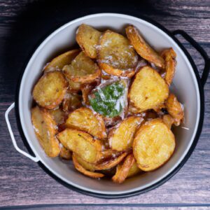 Esta imagem mostra uma porção de batatas assadas com crosta de parmesão, preparadas em uma airfryer. As batatas têm uma crosta dourada e crocante, com pedaços de queijo parmesão derretido. As batatas estão dispostas em um prato branco e são guarnecidas com salsa fresca.