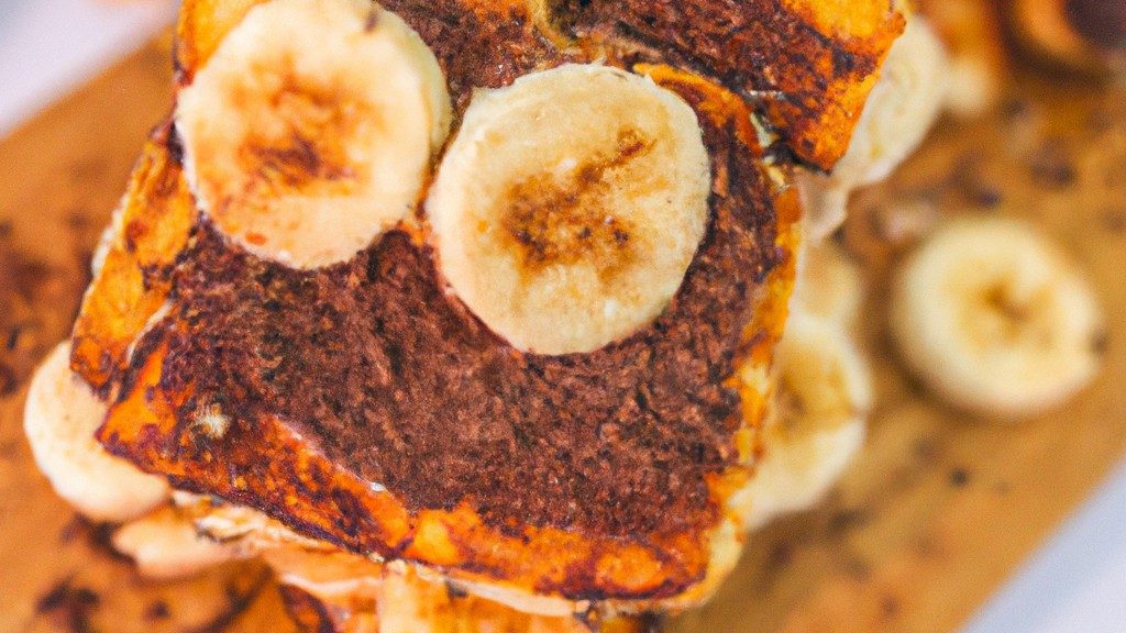 Imagem de uma torrada de banoffee feita na airfryer. Ela é composta por pão torrado coberto com bananas fatiadas, doce de leite e chantilly. A torrada é polvilhada com cacau em pó e apresentada em um prato branco.