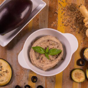 Imagem de uma porção de pasta de berinjela (babaganoush) assada em uma airfryer. A pasta é servida em um prato branco e decorada com folhas de hortelã e sementes de romã. O fundo é branco.