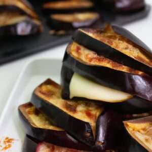 Imagem de uma torre de berinjela assada na airfryer, empilhada com camadas de queijo parmesão derretido, molho de tomate e folhas de manjericão. A torre está apoiada em uma superfície de madeira, com um fundo desfocado de utensílios de cozinha.