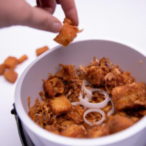 Imagem de cebolas crocantes fritas na Airfryer em um prato branco. As cebolas são cortadas em rodelas e cobertas com uma camada crocante dourada. A Airfryer é mostrada ao fundo, com a cesta aberta mostrando as cebolas cozidas.