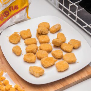 Imagem de nuggets congelados crocantes e dourados cozidos em uma Airfryer. Os nuggets são dispostos em uma cesta de arame da Airfryer e estão cobertos com gotas de óleo. O fundo da imagem é um fundo branco sólido.