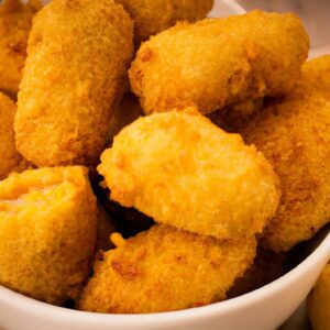 Uma foto de cima para baixo de um croquete de milho recheado com frango, coberto com uma crosta crocante e dourada, servido em uma cesta de vime em uma superfície de madeira.