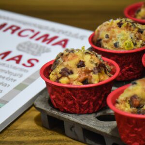 Imagem de muffins natalinos caseiros quentes e fumegantes feitos na Airfryer. Os muffins são decorados com glacê branco e vermelho e cobertos com granulado festivo. Eles estão dispostos em uma cesta forrada com papel manteiga, cercados por pinhas e azevinho.