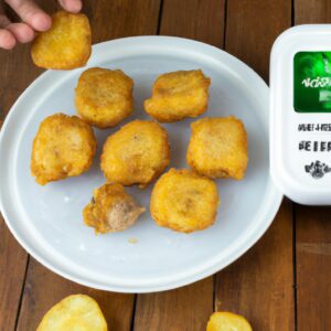 Imagem de nuggets de frango congelados dourados e crocantes servidos em uma tigela branca com um lado de molho de tomate. Os nuggets são empanados e têm um interior branco e macio. A tigela está sobre uma superfície de madeira com um pano de prato xadrez vermelho e branco ao lado.