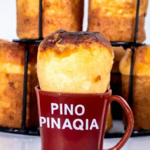 Imagem de um panetone de caneca na Airfryer. O panetone é marrom dourado e tem uma textura fofa, com uma cobertura de açúcar de confeiteiro polvilhada por cima. A caneca é branca e tem uma alça vermelha. Ao fundo, há uma Airfryer preta.
