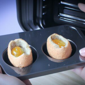 Imagem de um pão com ovo sendo preparado em uma airfryer. O pão é redondo e tem uma cavidade no centro, preenchida com um ovo. A airfryer é um pequeno eletrodoméstico com uma cesta de metal para cozinhar os alimentos.