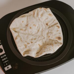 Imagem de um pão folha dourado cozido na Airfryer, uma fritadeira sem óleo. O pão tem um formato redondo e está ligeiramente achatado, com uma superfície texturizada e bolhas de ar visíveis. Ele está disposto sobre uma superfície branca com um fundo desfocado.